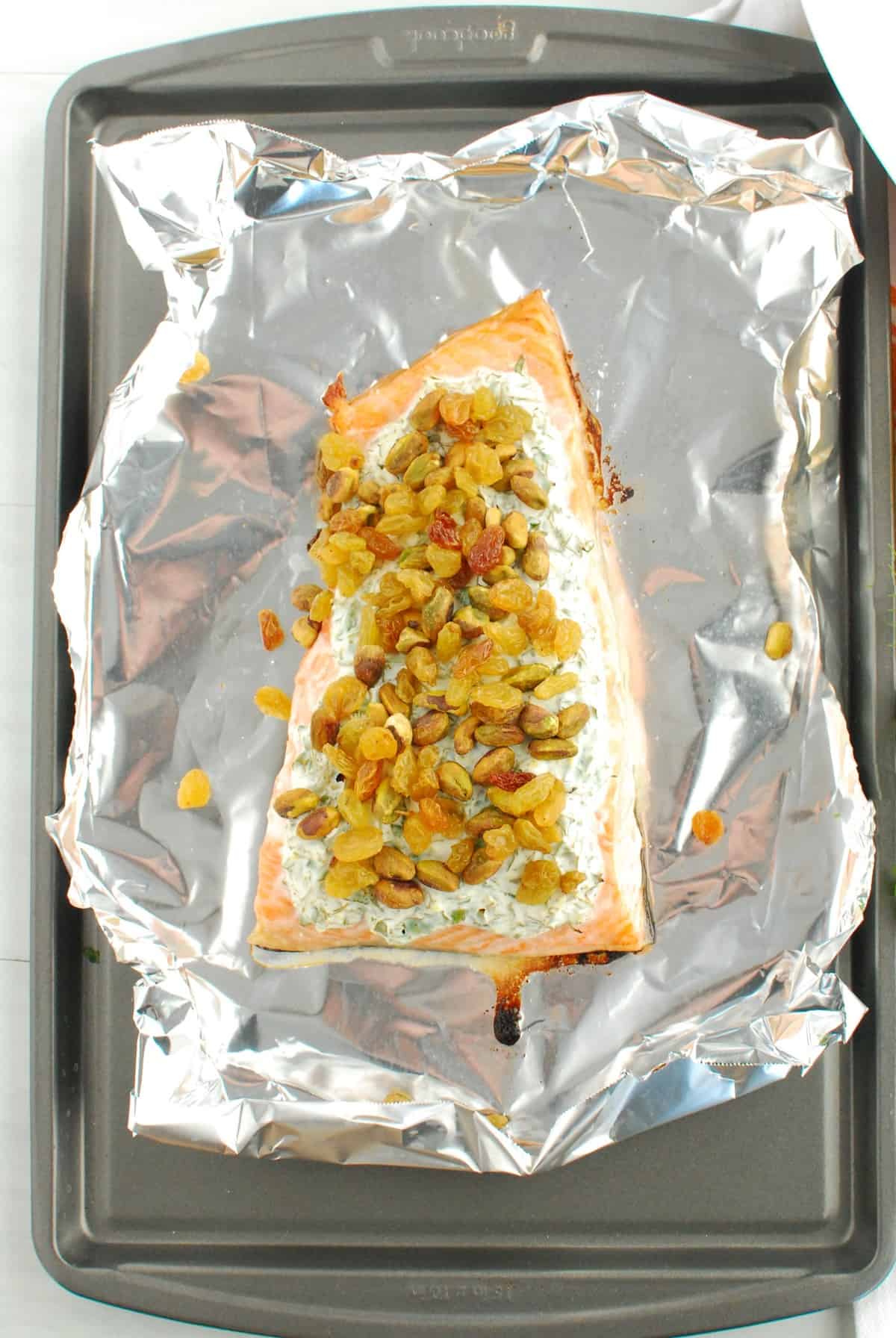 Baked salmon topped with herb yogurt, pistachios, and golden raisins on a foil-lined baking sheet.
