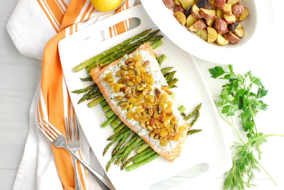A piece of salmon with herb yogurt, pistachios, and raisins, sitting over asparagus on a white platter, next to a napkin.
