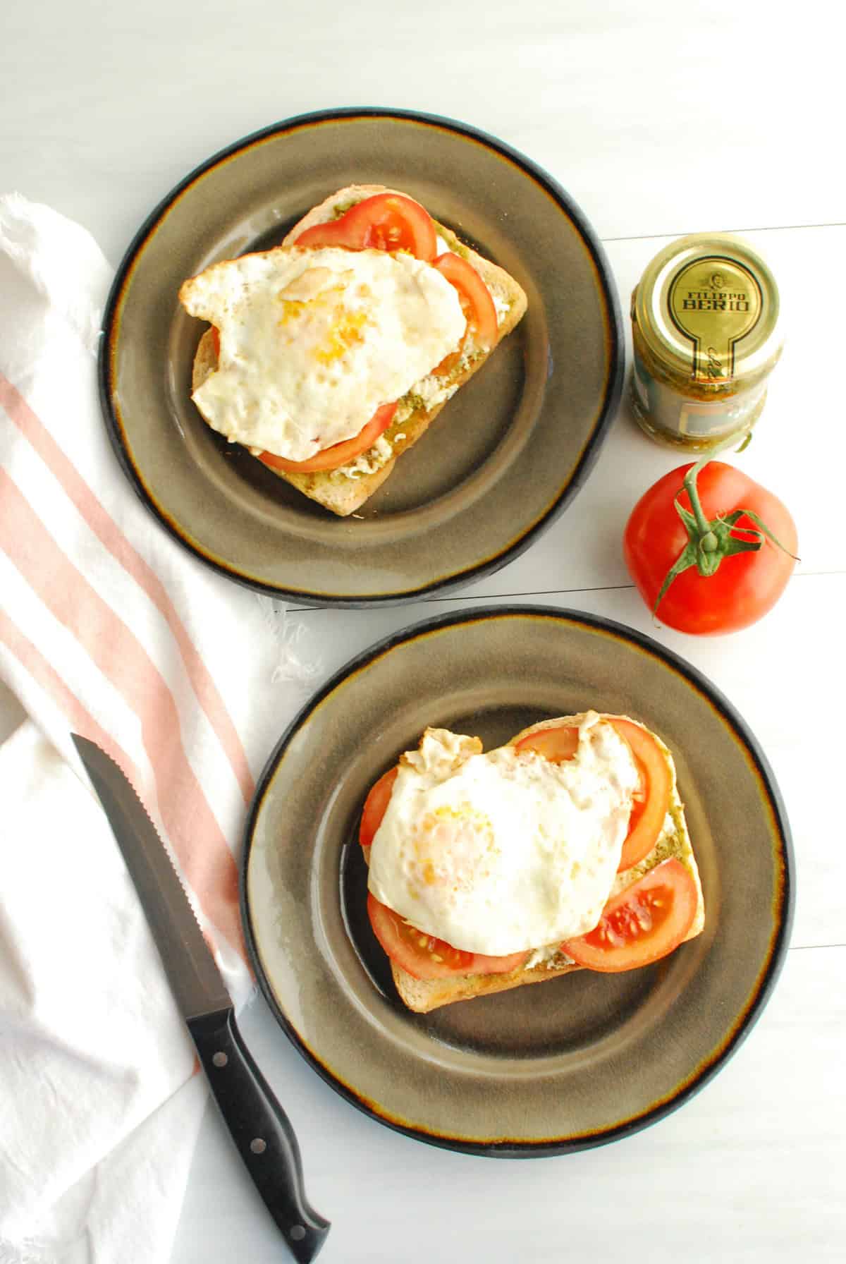 Overhead shot of two plates with pesto burrata toast next to a napkin, tomato, and jar of pesto.