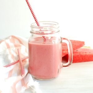 A glass with strawberry watermelon smoothie in it, next to a napkin and fresh watermelon slices.