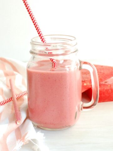 A glass with strawberry watermelon smoothie in it, next to a napkin and fresh watermelon slices.