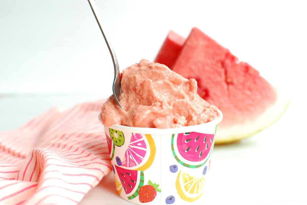 A cup of watermelon almond butter sherbet next to a napkin and freshly sliced watermelon.