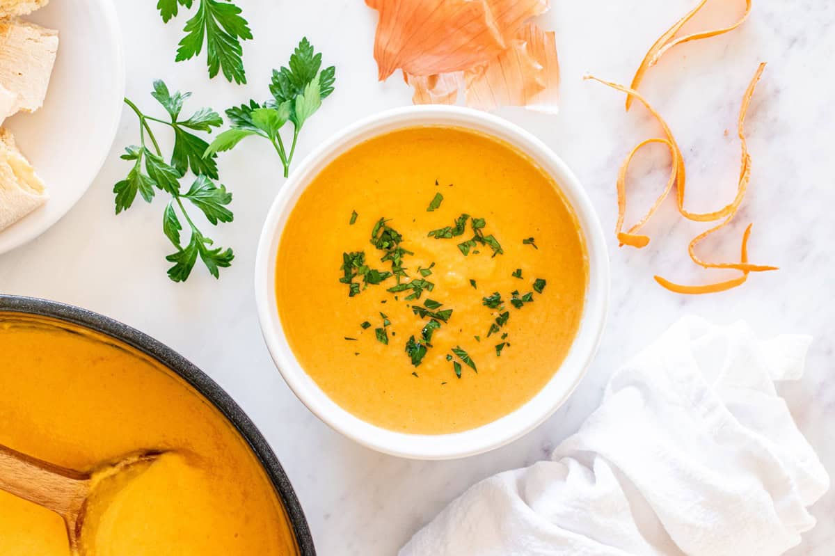 A white bowl filled with cauliflower carrot soup, next to some parsley, carrot peels, and a napkin.