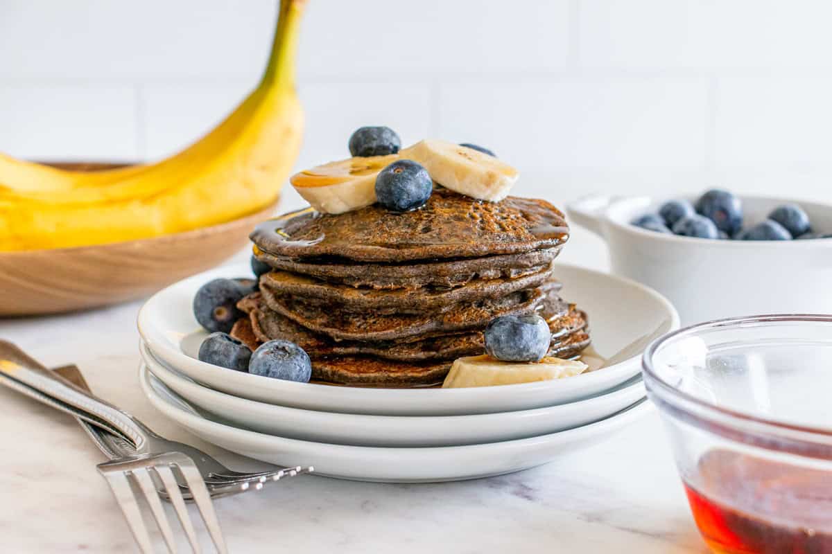 A stack of high fiber pancakes on a white plate topped with blueberries, banana, and maple syrup.