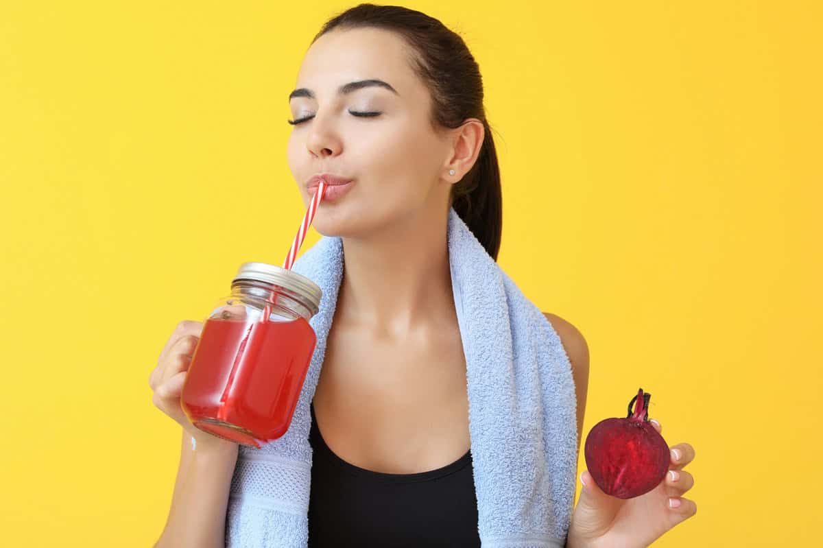 A woman drinking a glass of beet juice and holding a beet in her other hand.