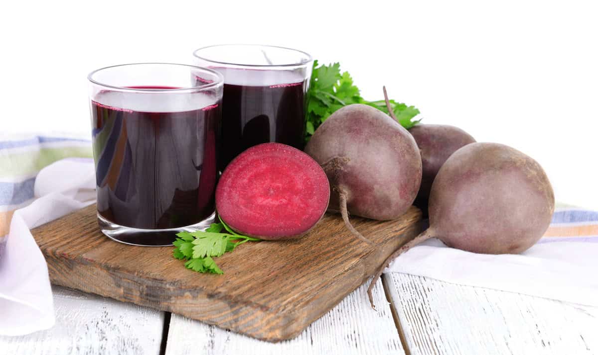 Two glasses of beet juice on a wooden cutting board next to fresh beets.