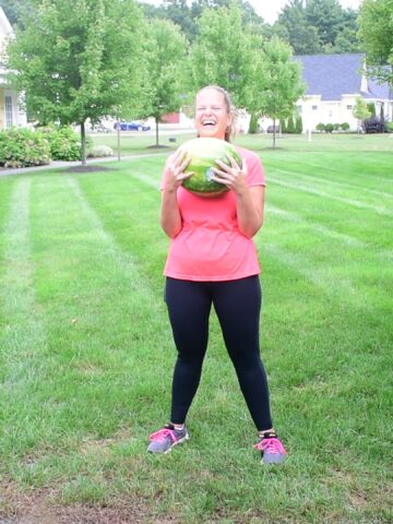 A woman holding a watermelon getting ready to workout with it, and laughing.