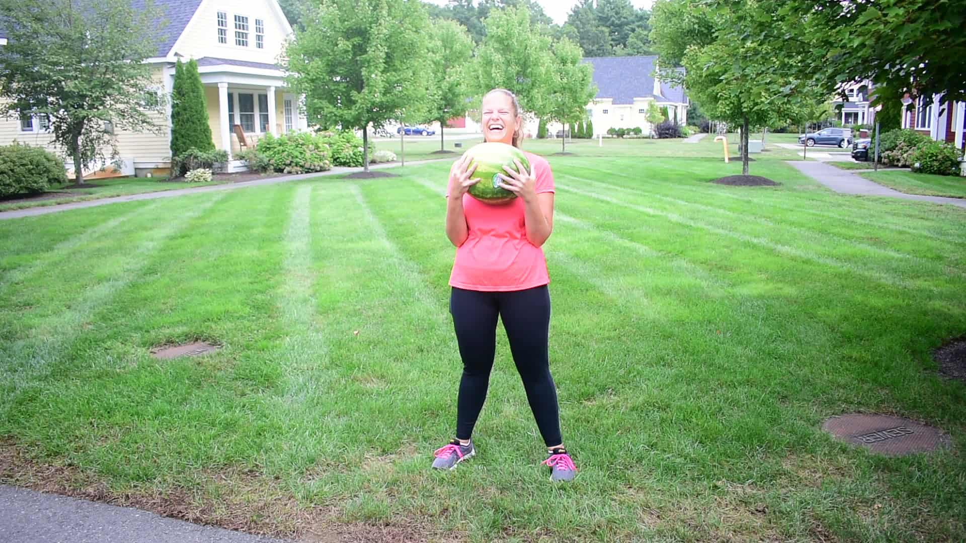 A woman holding a watermelon getting ready to workout with it, and laughing.