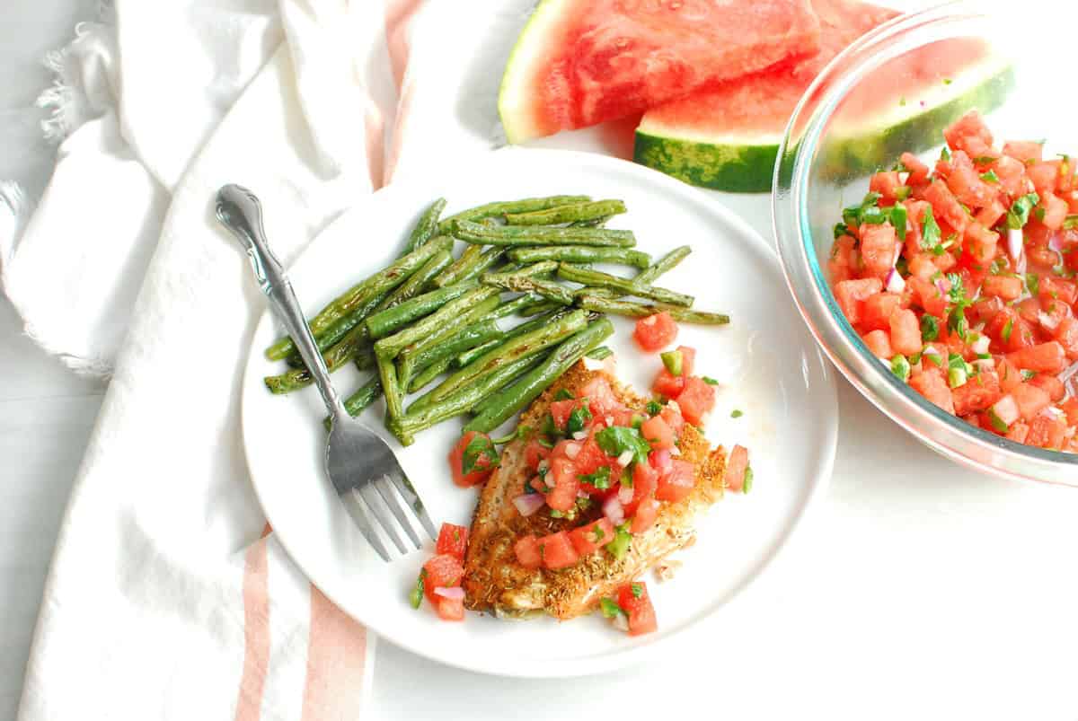 Salmon with watermelon salsa and roasted green beans on a white plate with a fork.