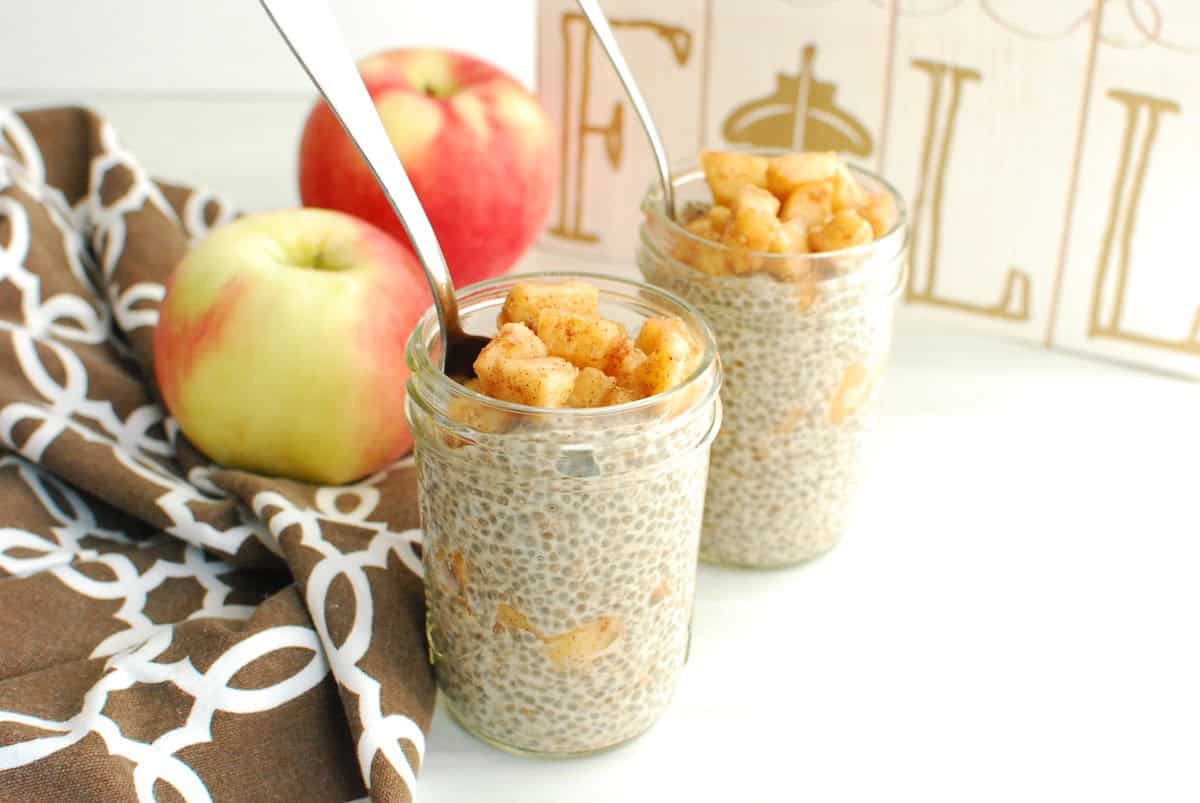 Two mason jars of apple chia pudding next to a napkin and some fresh apples.