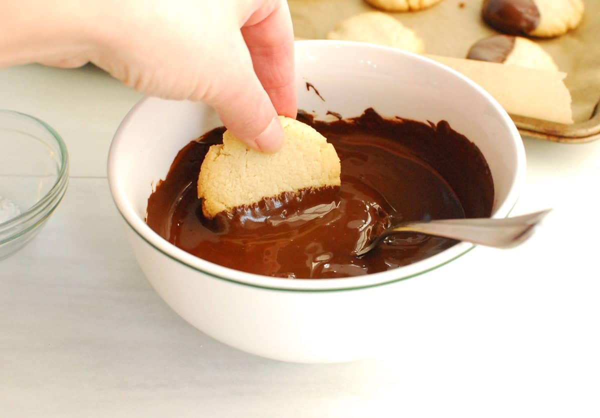 A woman's hand holding a cookie and dipping it in a bowl of melted chocolate.