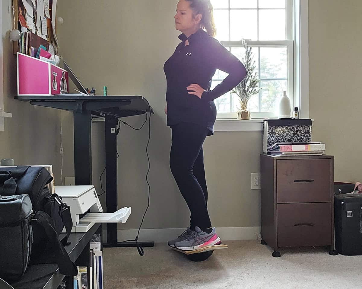 A woman balancing on a Whirly Board at a standing desk.