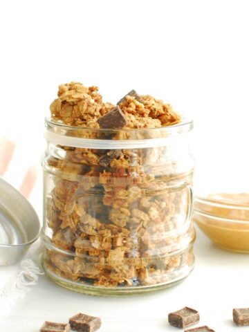A jar filled with peanut butter chocolate protein granola, next to some chocolate chips and peanut butter.