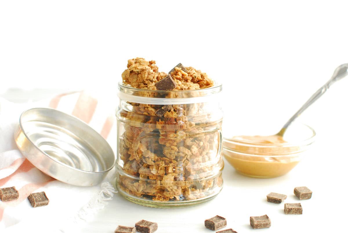 A jar filled with peanut butter chocolate protein granola, next to some chocolate chips and peanut butter.