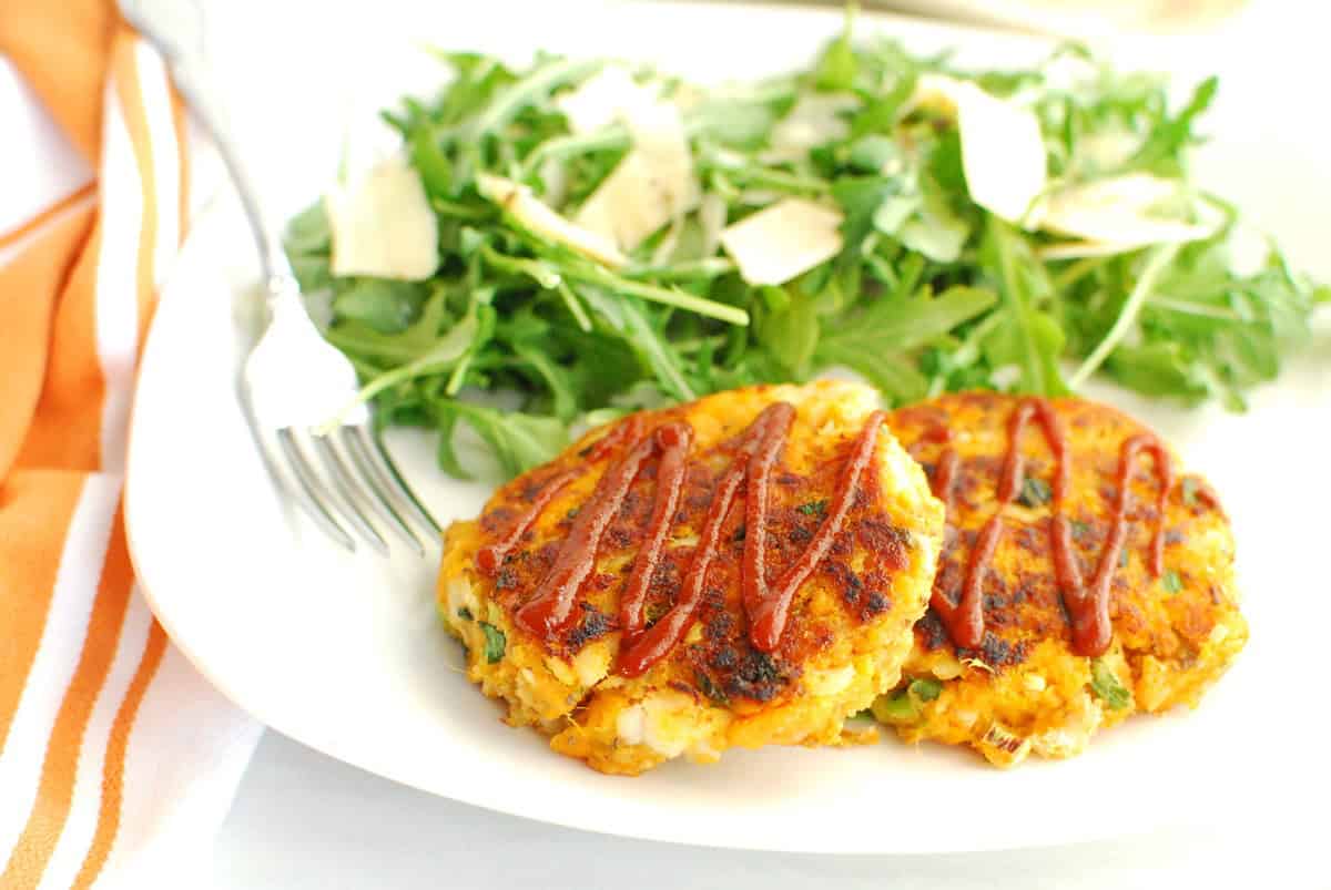 Two sweetpotato fish cakes on a plate topped with sriracha next to an arugula salad.