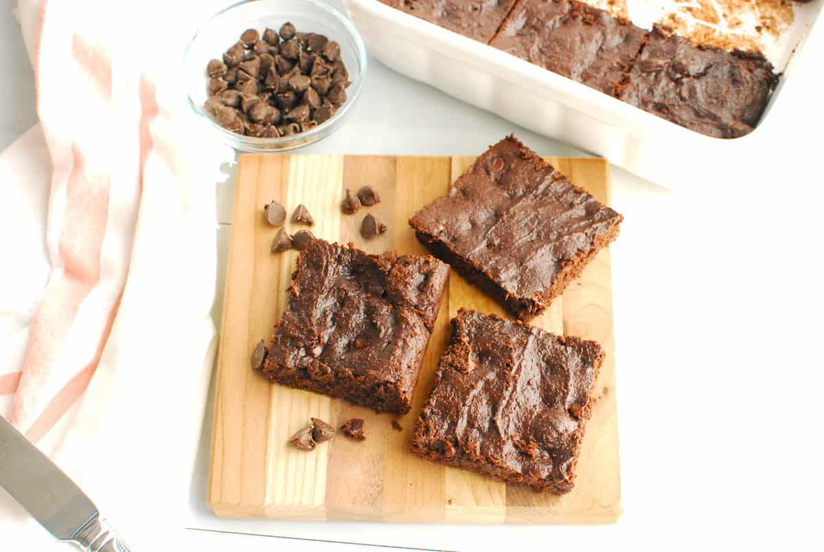 Three cottage cheese brownies on a cutting board next to some chocolate chips.
