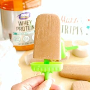 A peanut butter chocolate protein ice pop with a canister of protein powder in the background.