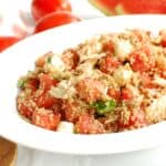 A bowl of watermelon quinoa salad next to some fresh watermelon and tomatoes.