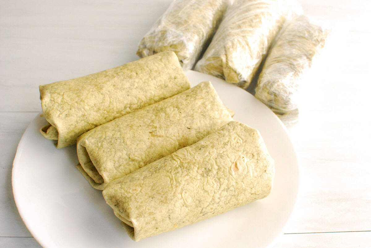 Several breakfast burritos on a plate, with a few rolled in plastic wrap in the background.