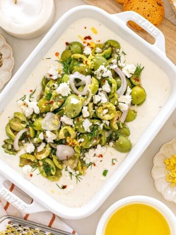 A white dish filled with whipped feta dip with olives, surrounded by some of the ingredients.