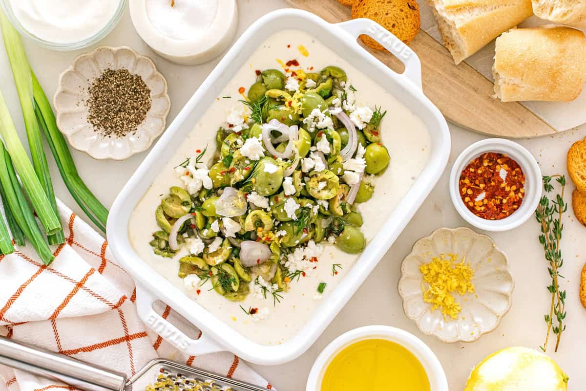A white dish filled with whipped feta dip with olives, surrounded by some of the ingredients.