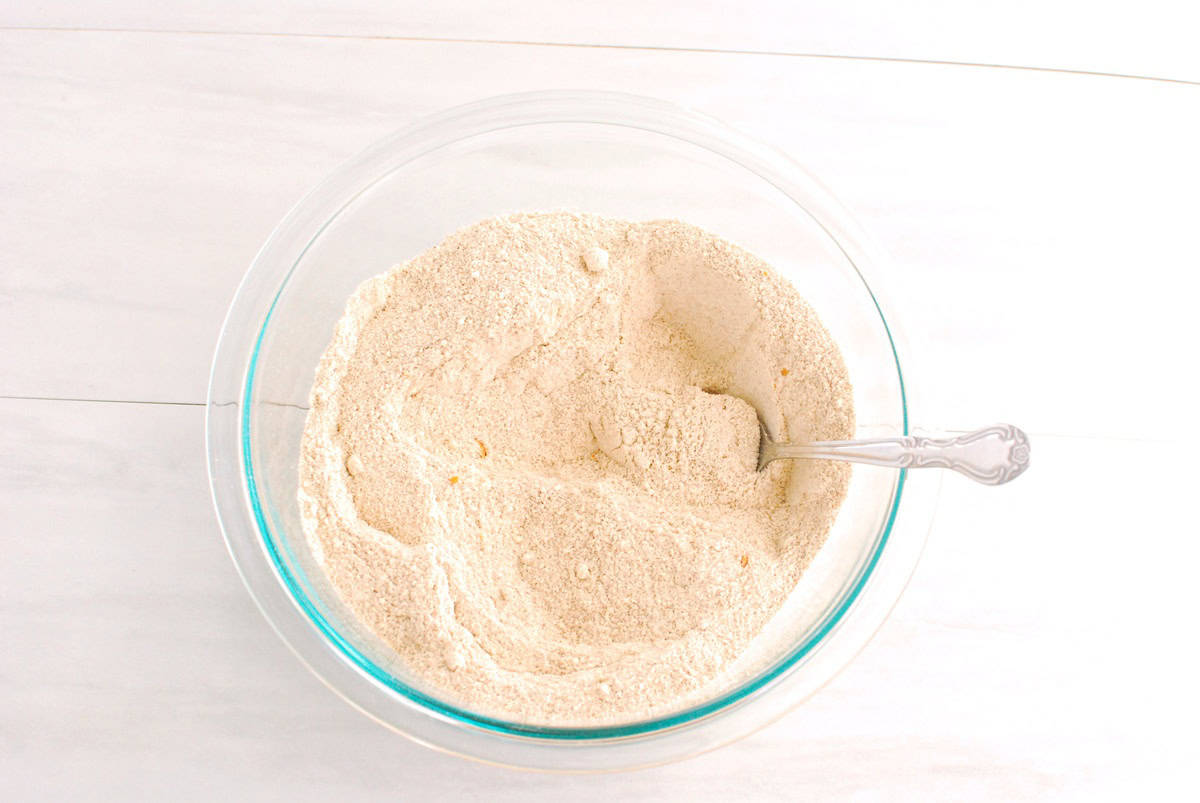 Protein powder, whole wheat flour, almond flour, orange zest, cinnamon, baking powder, baking soda, and salt mixed together in a glass bowl.