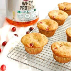 Cranberry protein muffins on a cooling rack next to a napkin, fresh cranberries, and whey protein powder.