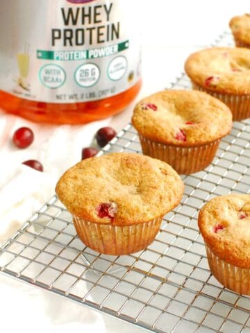 Cranberry protein muffins on a cooling rack next to a napkin, fresh cranberries, and whey protein powder.