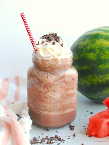 A mason jar of watermelon frozen hot chocolate next to some fresh watermelon and chocolate shavings.