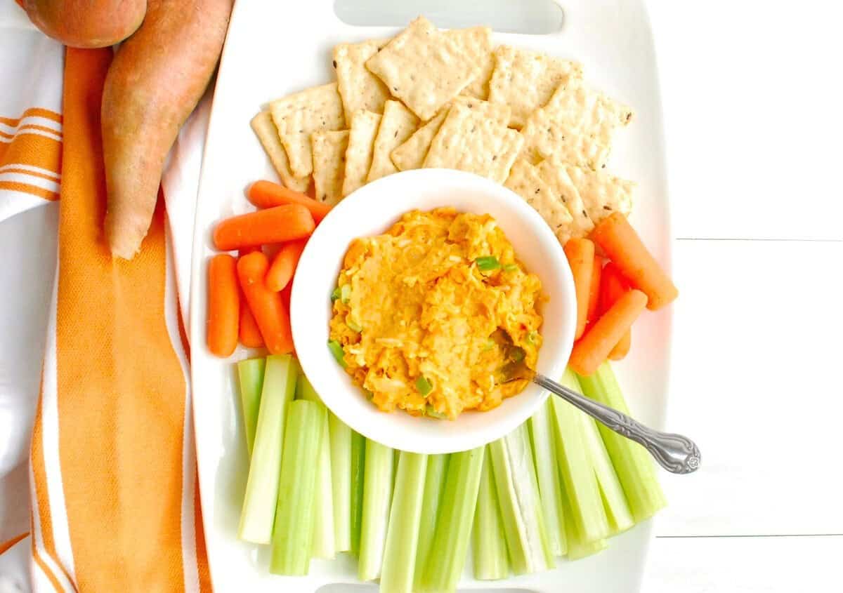 Vegetarian buffalo dip in a bowl on a platter with celery, carrots, and crackers.