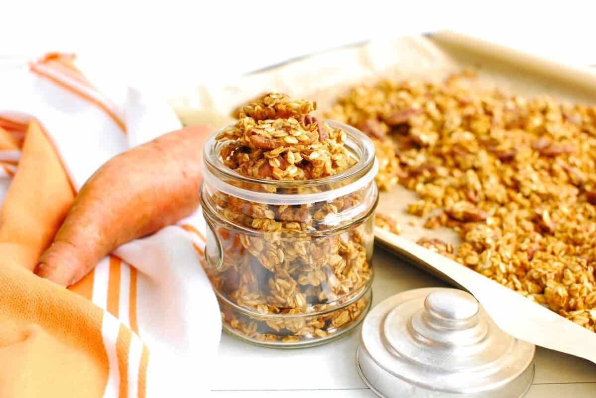 A jar of sweetpotato granola next to a baking sheet and fresh sweetpotato.