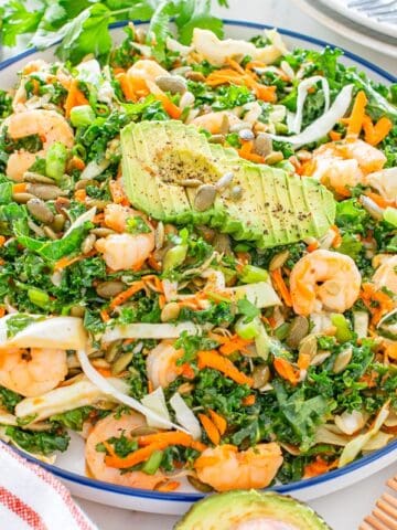 A bowl of kale salad with shrimp, avocado, pepitas, and other ingredients.