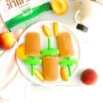 Three peach crisp ice pops next to fresh sliced peaches, oat milk powder, and maple syrup.