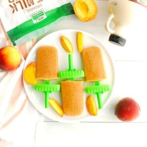 Three peach crisp ice pops next to fresh sliced peaches, oat milk powder, and maple syrup.