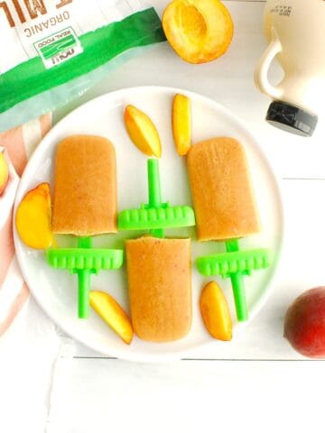 Three peach crisp ice pops next to fresh sliced peaches, oat milk powder, and maple syrup.