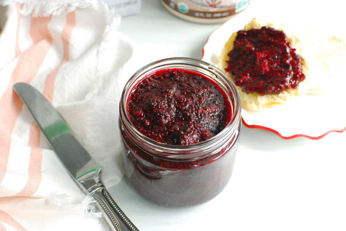 A jar of blackberry chia seed jam next to a knife and an English muffin topped with jam.