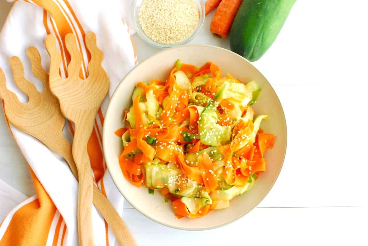A bowl filled with sesame carrot cucumber salad next to wooden serving tongs.