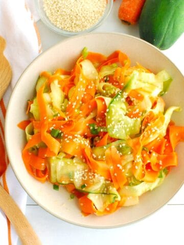 An overhead view of a bowl of sesame cucumber carrot salad next to tongs and a napkin.