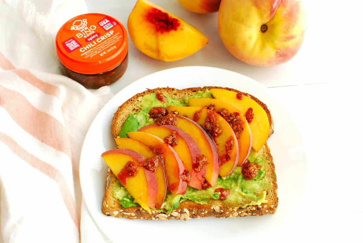 A slice of avocado peach toast on a plate next to some fresh peaches and a container of chili crisp.