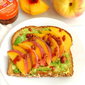 A slice of peach avocado toast topped with chili crisp on a white plate.
