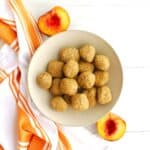 A bowl filled with peach pie energy balls next to a fresh sliced peach.