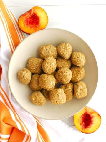 A bowl filled with peach pie energy balls next to a fresh sliced peach.