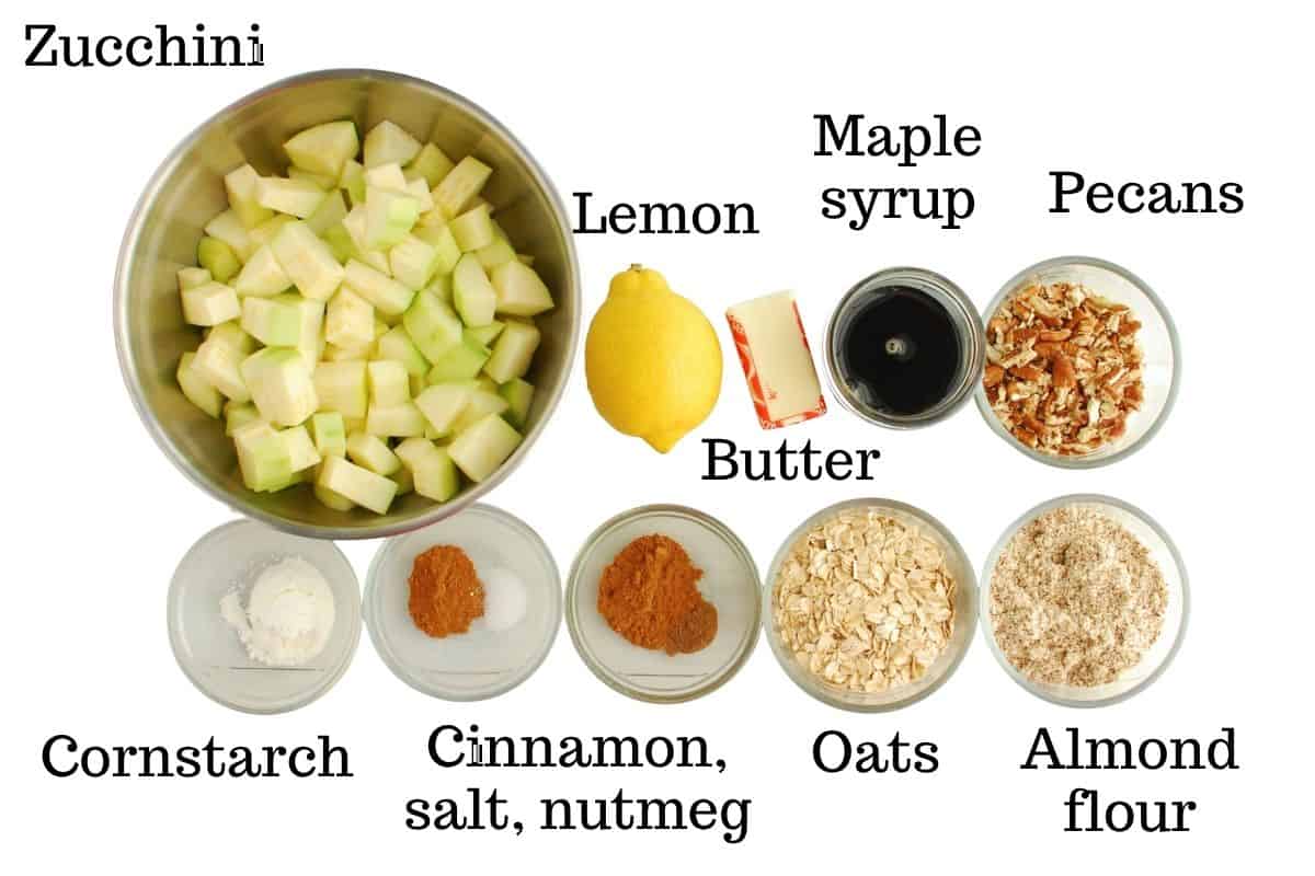Zucchini, lemon, butter, maple syrup, pecans, cinnamon, salt, nutmeg, cornstarch, oats, and almond flour, all on a white backdrop.