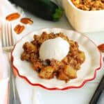 A plate of zucchini crisp topped with ice cream next to a fork and the baking dish.