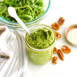 A jar of arugula pesto next to some fresh arugula and pecans on a table.